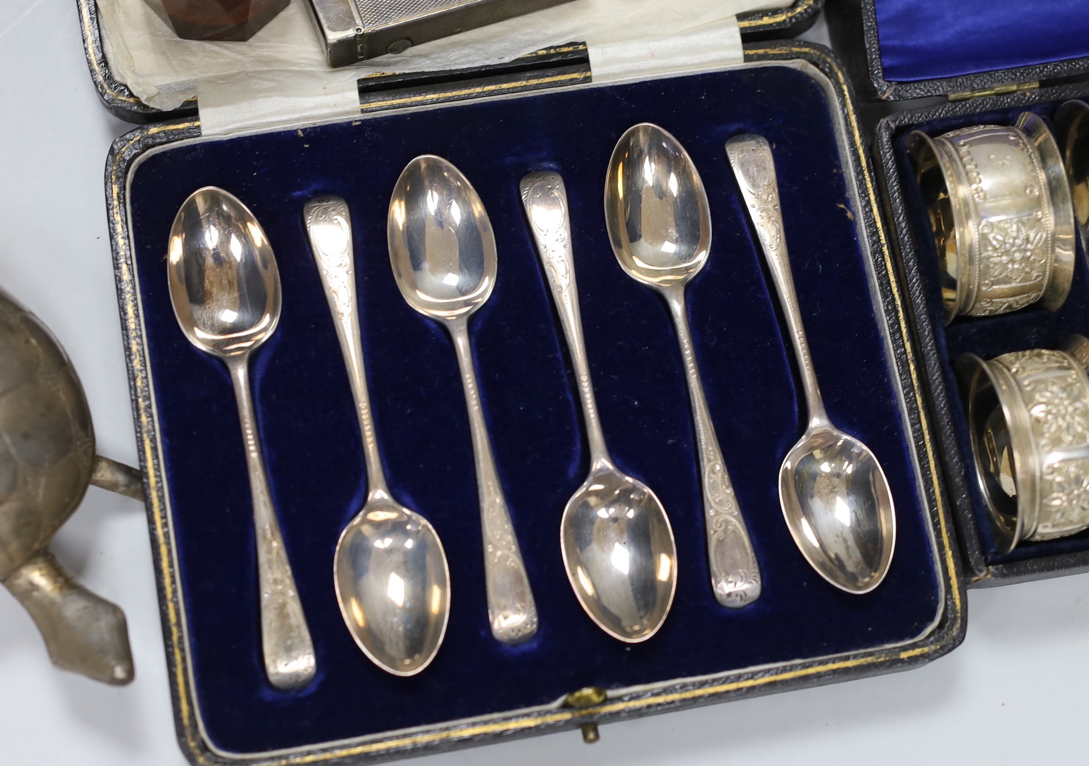 A cased set of six George V silver teaspoons, a white metal mounted glass scent bottle, a Dunhill lighter, a plated vesta, a white metal tortoise and a cased set of six plated napkin rings.